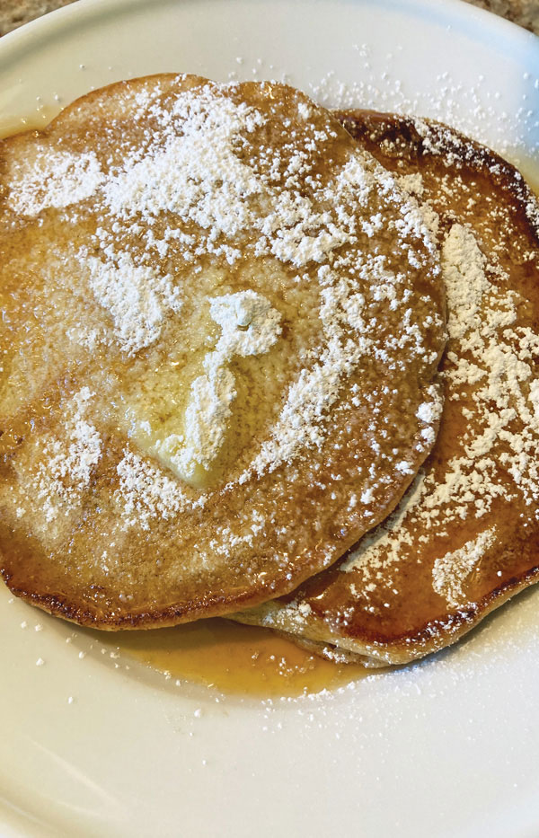 New Fashioned Pancakes shown with maple syrup and powdered sugar on a plate.